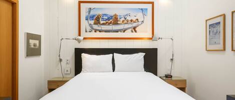 Une chambre d’hôtel avec un grand lit, deux tables de chevet, un tableau représentant un chien sur un bateau, accroché au mur, et une lampe fixée au mur.