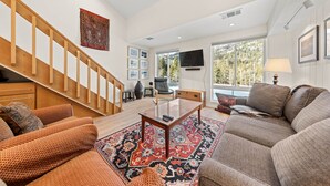 Un salon spacieux avec un escalier en bois, une grande fenêtre donnant sur la forêt, un téléviseur à écran plat et un coin salon confortable agrémenté de coussins à motifs.