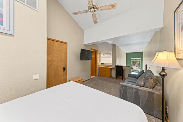 A hotel room with a bed, a desk, a chair, a sofa, a ceiling fan, a TV, and a lamp.