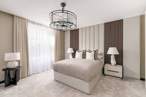 A modern bedroom with a large bed, two bedside tables with lamps, a chandelier, and a view of the outdoors through sheer curtains.