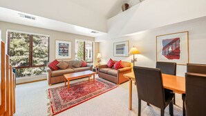 A living room with two sofas, a coffee table, a dining table with chairs, and large windows with blinds.