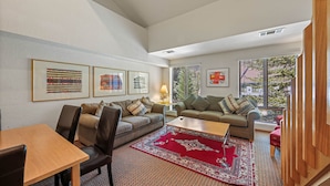 A living room with a sofa, a coffee table, and a rug. There are framed pictures on the wall and a window with blinds. - 305 | Pool View A/C Loft!  - Tubs, Sauna & Fun (Olympic Valley)