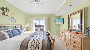 A bedroom with a bed, a dresser, a ceiling fan, a window with curtains, and a city view.