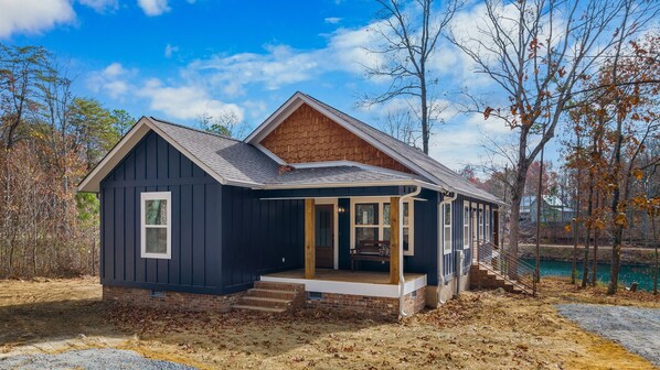 A small, dark-colored cabin with a porch and a brick foundation, surrounded by trees and a body of water.