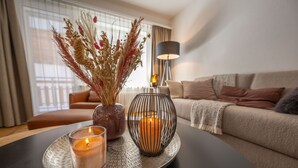 Een gezellige woonkamer met een ronde tafel waarop kaarsen en een vaas met gedroogde bloemen staan. Een bank en een lamp staan op de achtergrond.