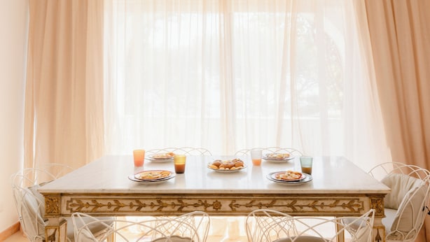 Une table à manger dressée pour le petit-déjeuner, avec des assiettes remplies de nourriture et des boissons, entourée de chaises blanches.