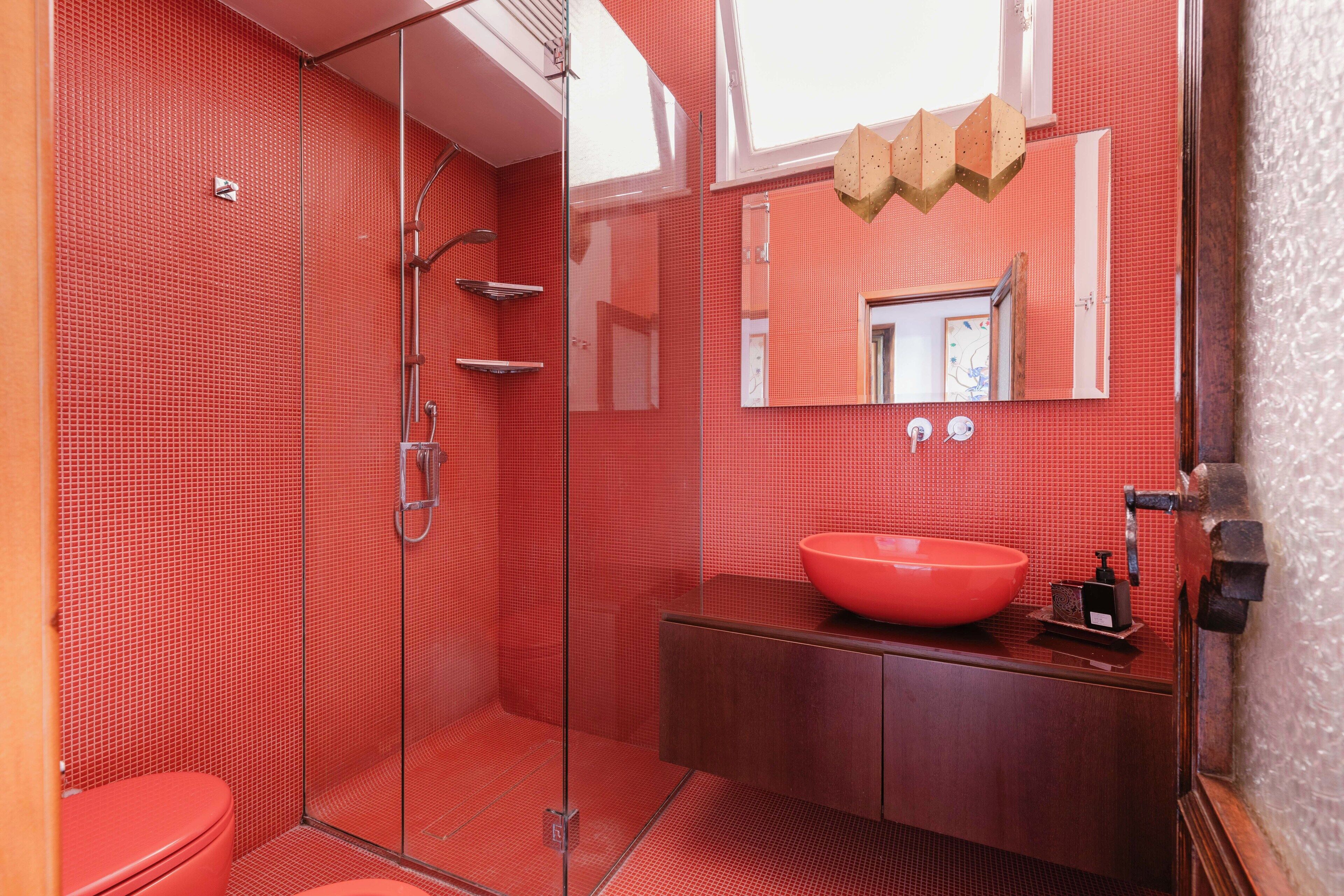 A modern bathroom with a red tiled wall, a glass shower enclosure, a wooden vanity with a red sink, and a large mirror.