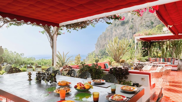 Un espace de restauration en plein air avec une table dressée pour un repas, entourée de chaises et surmontée d’un auvent rouge.