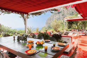 Un espace de restauration en plein air avec une table dressée pour un repas, entourée de chaises et surmontée d’un auvent rouge.