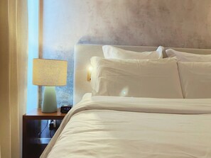 Een net opgemaakt bed met witte lakens, een bedlampje en een digitale klok op een nachtkastje van hout.