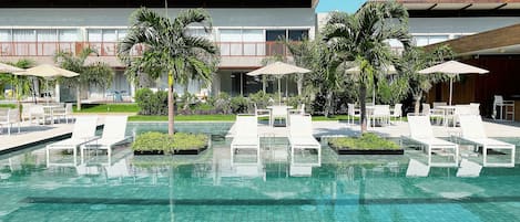 Piscina con sillones de descanso y sombrillas, rodeada de palmeras y un edificio de diseño moderno.