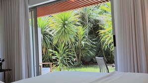 A bedroom with a large bed, a view of a garden with various plants, and a balcony with a wooden roof. - Flat Ilê de Pipa Jardim - By Almare (Tibau do Sul)