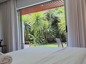 A bedroom with a large bed, a view of a garden with various plants, and a balcony with a wooden roof.