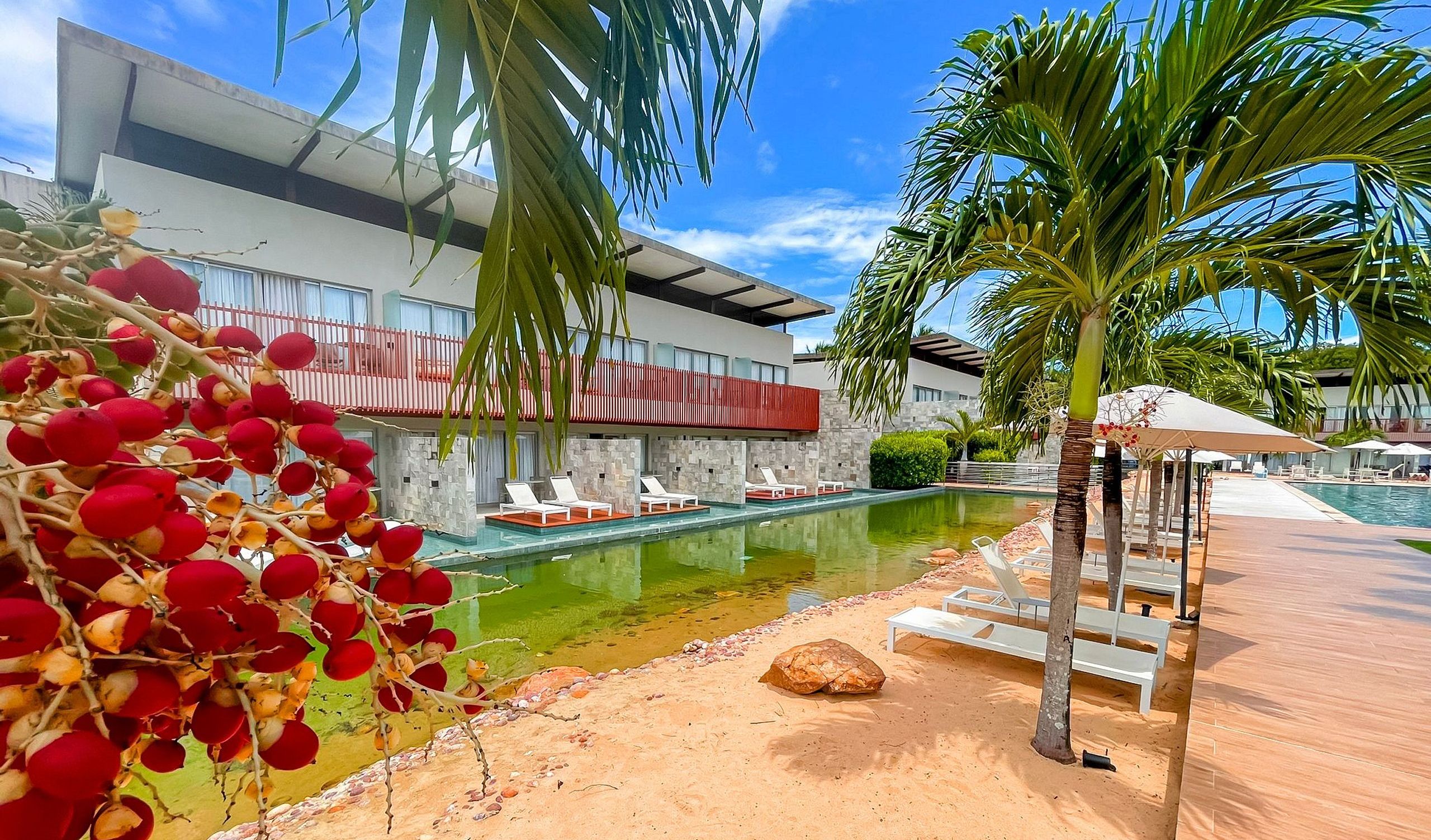A poolside area with lounge chairs, a palm tree, and a modern building with a red balcony.