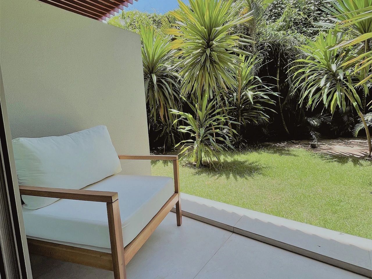 Uma varanda com uma cadeira de madeira, um travesseiro branco e vista para um jardim com diversas plantas.