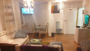 Uma sala de estar com sofá, uma mesa de jantar de vidro e cadeiras, televisão e uma pequena cozinha compacta.
