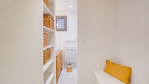 Un couloir étroit, doté d’étagères et de paniers blancs, mène à un banc blanc agrémenté d’un coussin jaune.