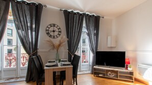 A living room with a wooden table, two chairs, a TV on a stand, and a large window with curtains.