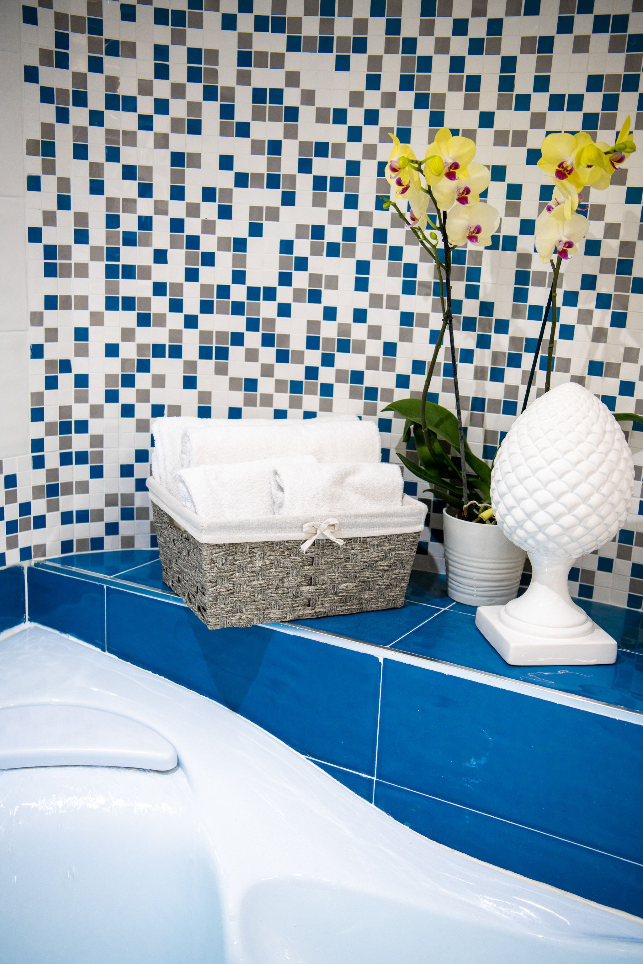 Une salle de bain avec un sol carrelé de carreaux bleus, une baignoire blanche, un panier en osier rempli de serviettes, une plante en pot et un objet décoratif blanc.