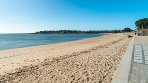 Ein Strand mit blauem Himmel, sandigem Ufer und einem Gebäude, von dem aus ein Weg zum Wasser führt.