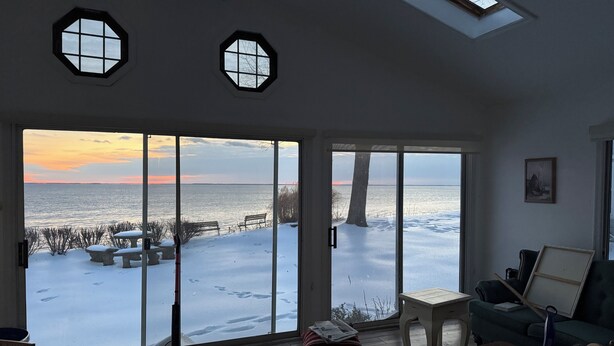 A room with large windows overlooking a snowy landscape and a body of water at sunset.