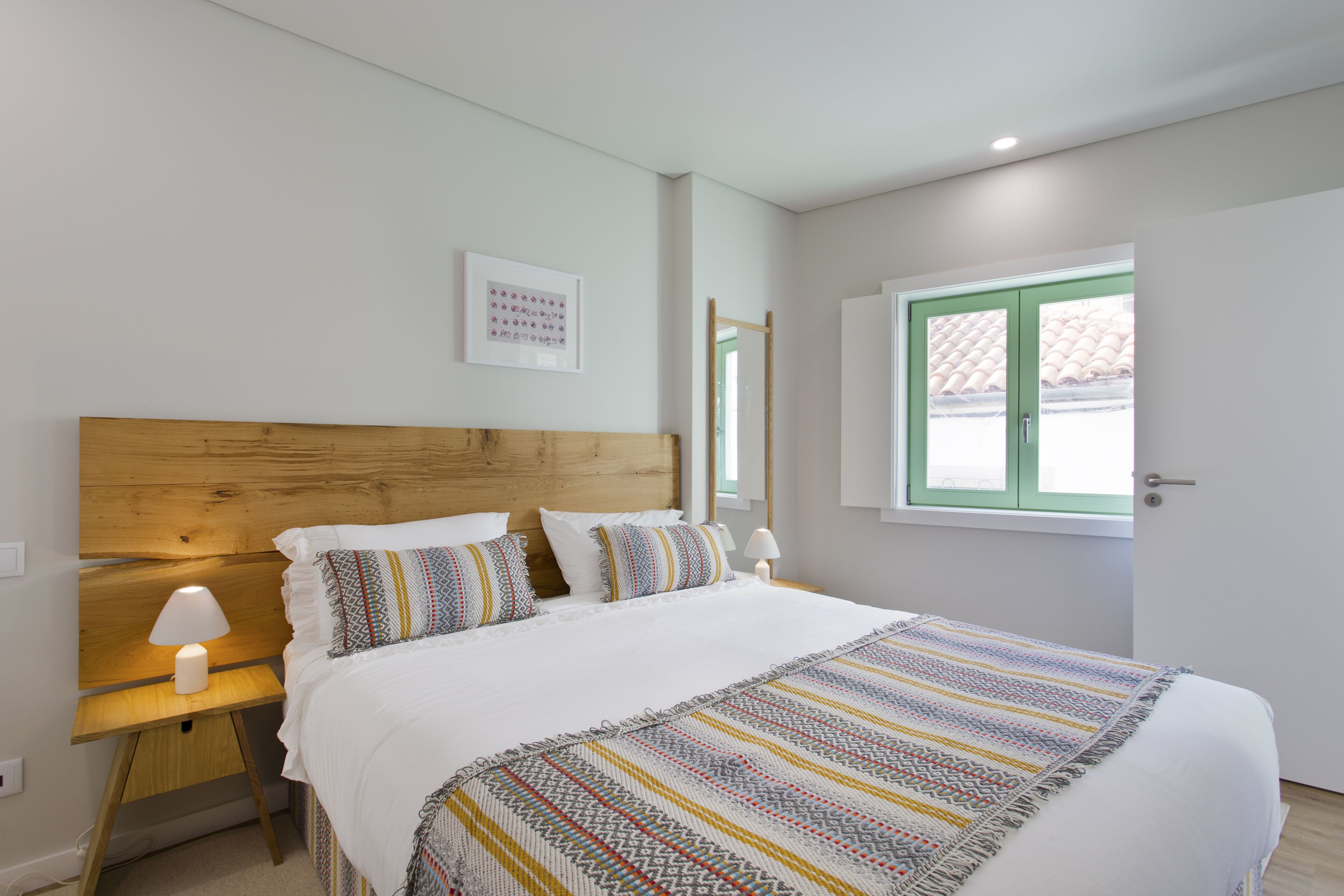A bedroom with a wooden headboard, white bedding, a patterned throw blanket, and a bedside lamp.