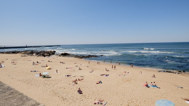 Una amplia playa de arena con gente tomando el sol y nadando, un rompeolas rocoso y el océano al fondo.