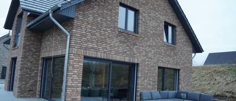 Une maison moderne avec une façade en briques, de grandes portes-fenêtres coulissantes et un patio aménagé avec du mobilier d’extérieur.