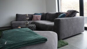 A modern living room with a grey sectional sofa, a small side table, and a green throw blanket on a grey ottoman. - Ferienhaus in Lüneburg (Westergellersen)