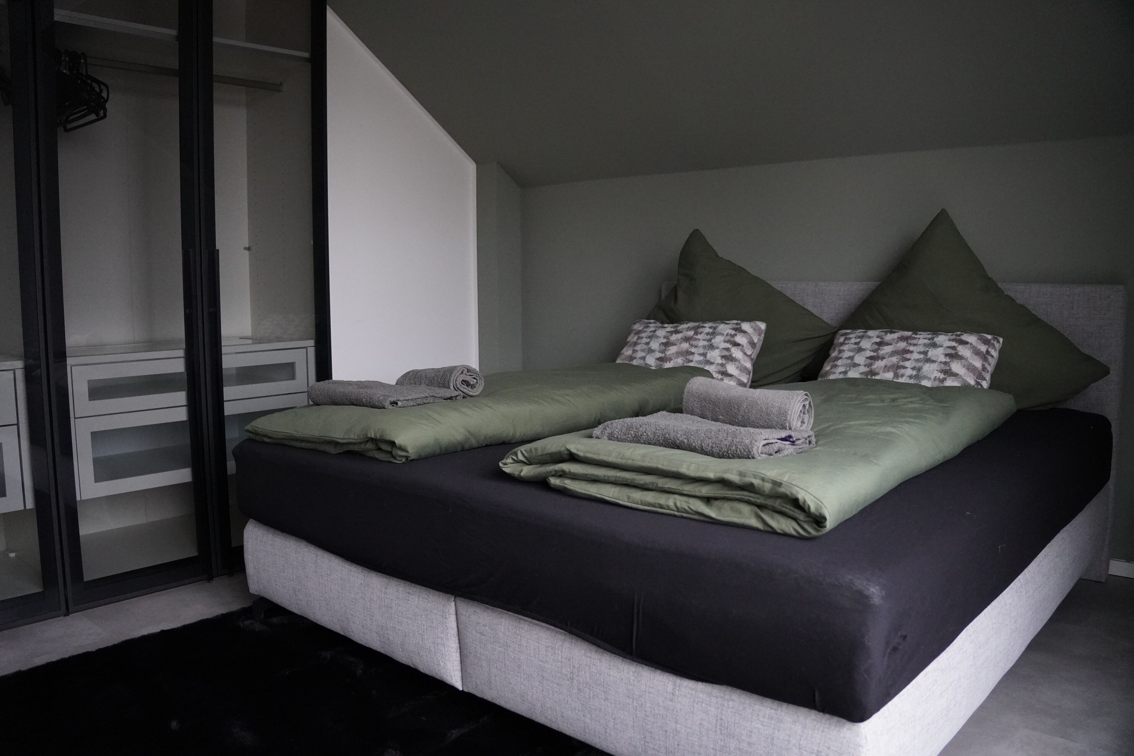 A modern bedroom with a bed, grey and green bedding, and a large window.