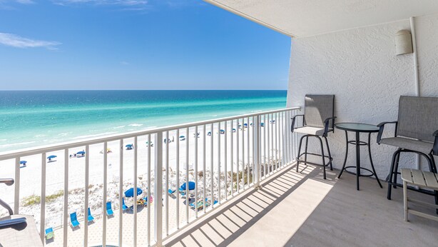A balcony with a view of the beach and ocean. There are chairs and tables on the balcony.