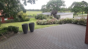 A paved patio area with two large planters, a grassy lawn, and a white building in the background. - Liebevoll Saniertes Ferienhaus in Ruhiger Warftlage in Dagebüll-kirche (Dagebüll-Kirche)