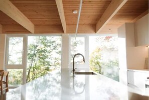 A modern kitchen with a large island, wooden ceiling beams, and large windows offering a view of trees. - Tree house Chez Leon (petite rivière saint françois)