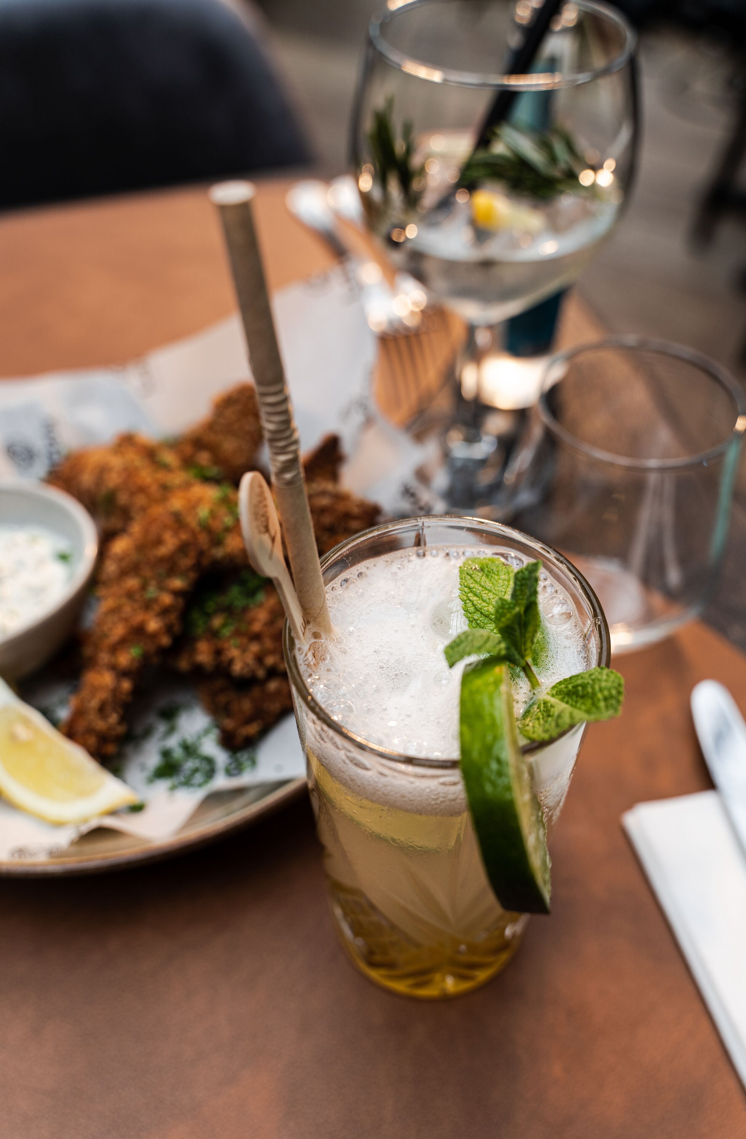 Une table avec un verre de bière garni d’une tranche de citron vert et de menthe, une assiette de nourriture frite et un verre d’eau avec une paille.