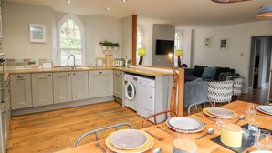 A modern kitchen with a built-in washing machine, a dining area with a wooden table set for four, and a living area with a sofa and a TV. - Upper Vestry (Llandysul)