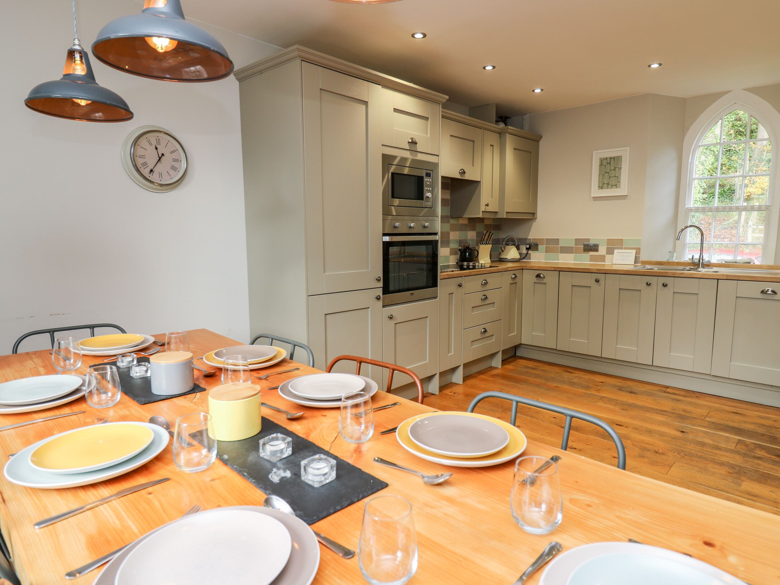 A modern kitchen with light-colored cabinets, a wooden dining table set for four, and a large window providing natural light.