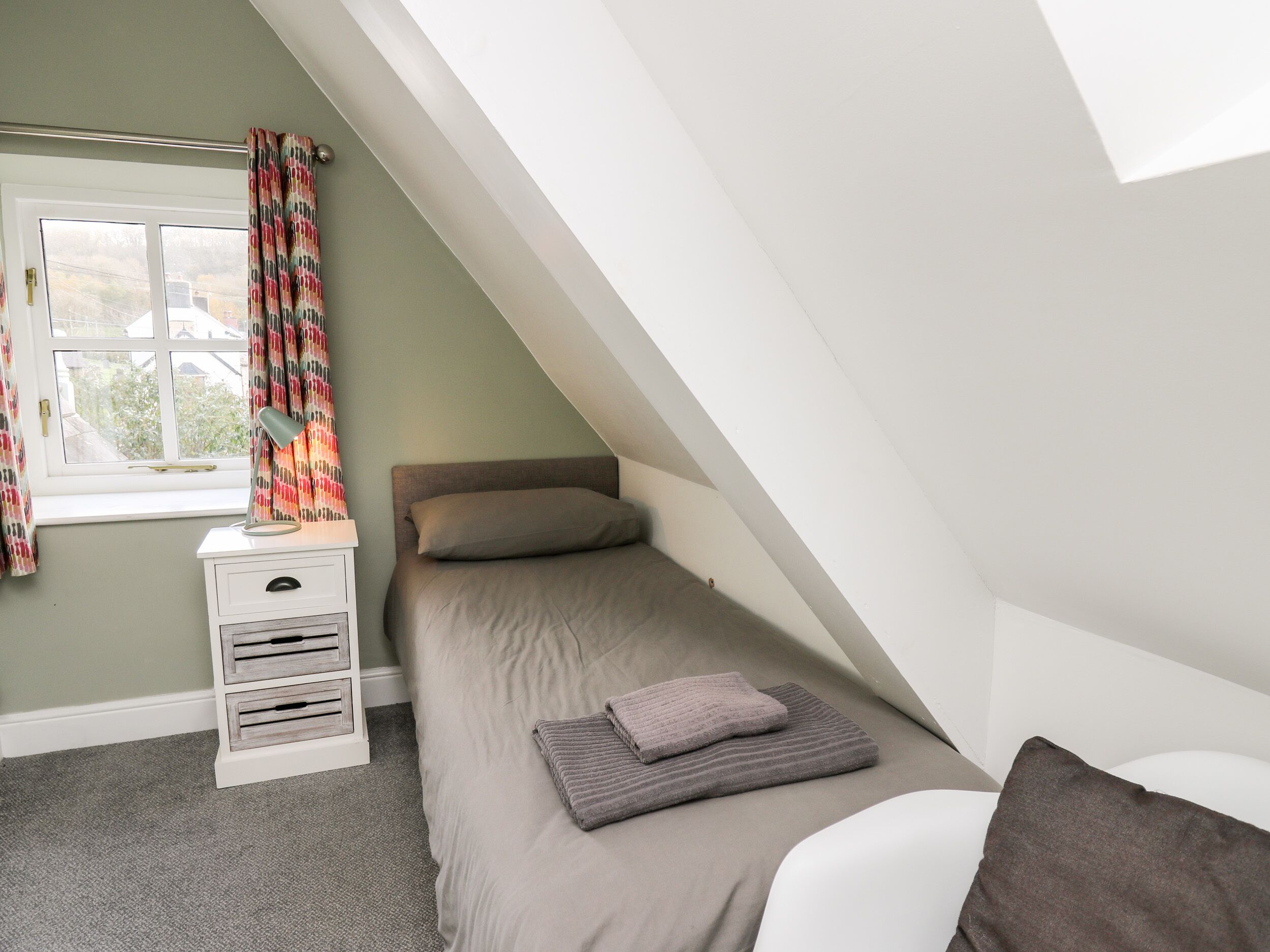 Ein kleines Zimmer mit einem Einzelbett, geneigter Decke, einem fenster mit Vorhängen, einem Nachttisch mit Schubladen und einer Nachttischlampe.