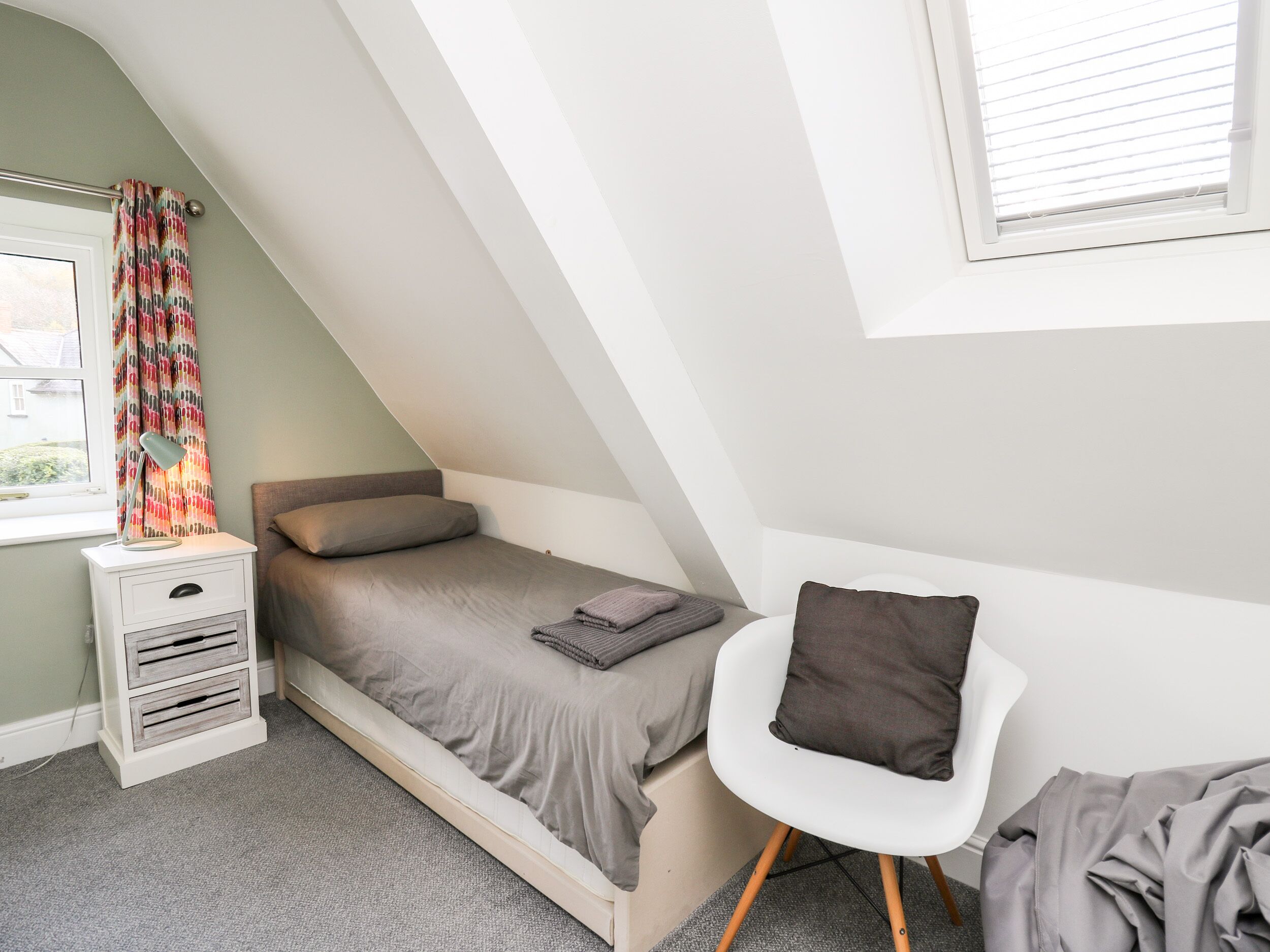 A small, neatly arranged bedroom with a single bed, a bedside table with drawers, a chair, and a window with blinds.
