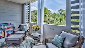 Uma área de estar coberta ao ar livre com móveis de vime, uma mesa pequena com um vaso de flores e vista para o verde através de grandes janelas.