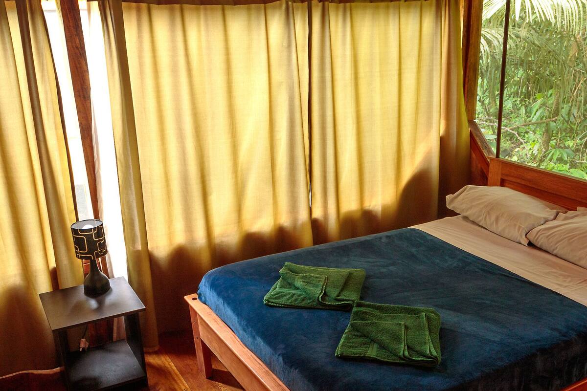 A bedroom with a bed, a nightstand, and a window with a view of greenery.