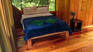 A wooden bed with a blue blanket and two green towels, a nightstand with a lamp, and a wooden floor.