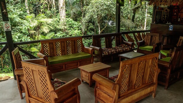 A wooden outdoor seating area with benches and chairs, surrounded by lush greenery.