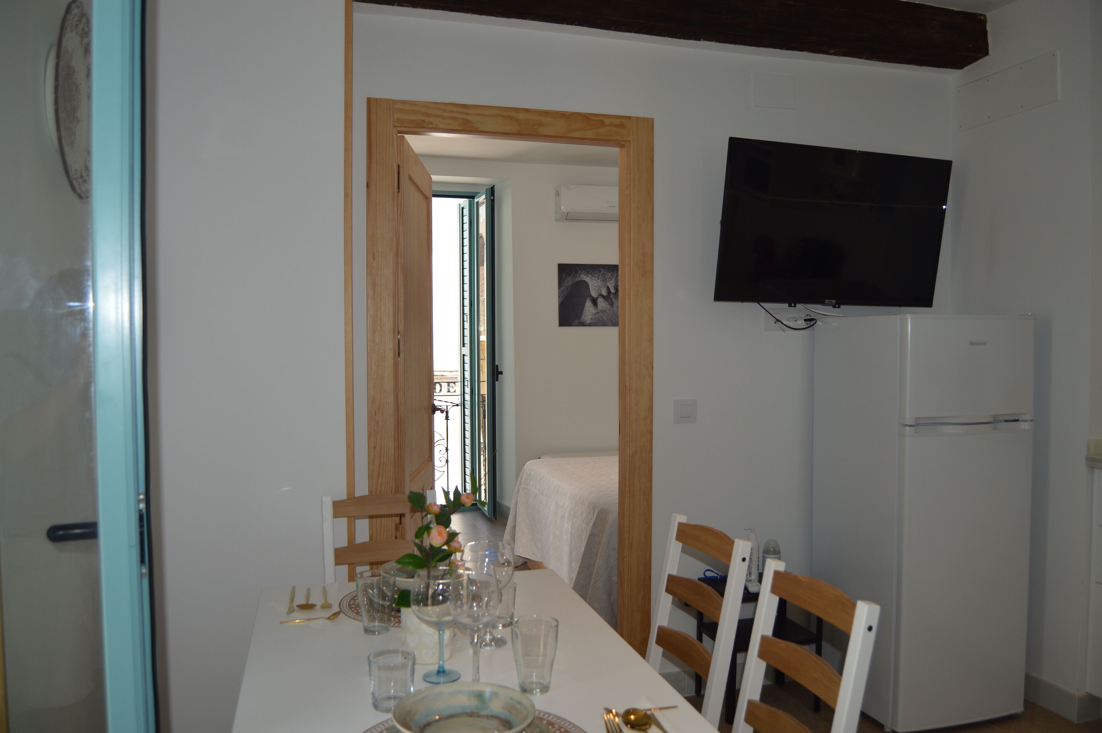 A dining area with a table set for four, a television mounted on the wall, and a refrigerator to the right.
