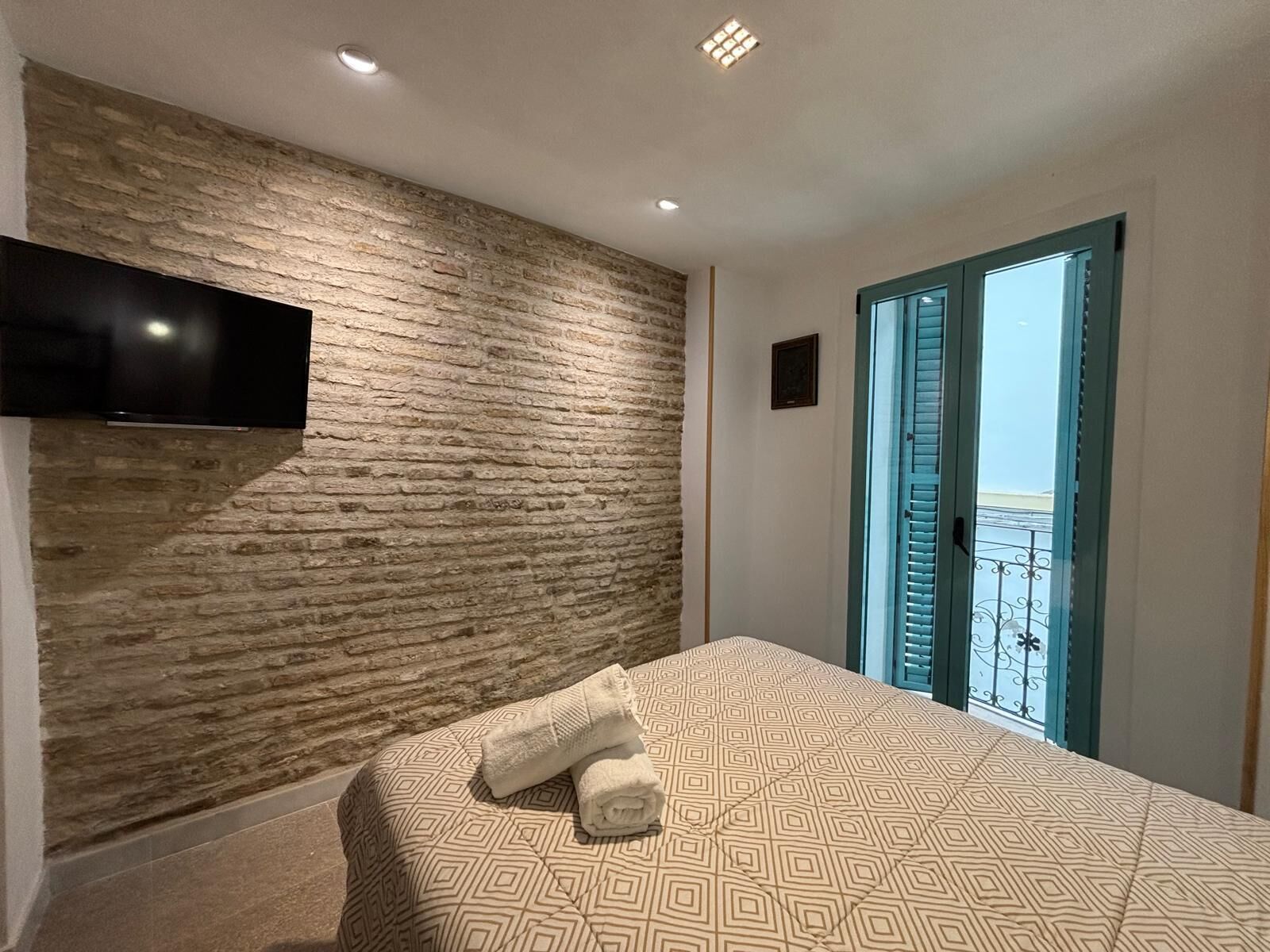A bedroom with a brick-patterned headboard, a flat-screen TV mounted on the wall, a bed with a patterned bedspread, and a door leading to a balcony with a view.