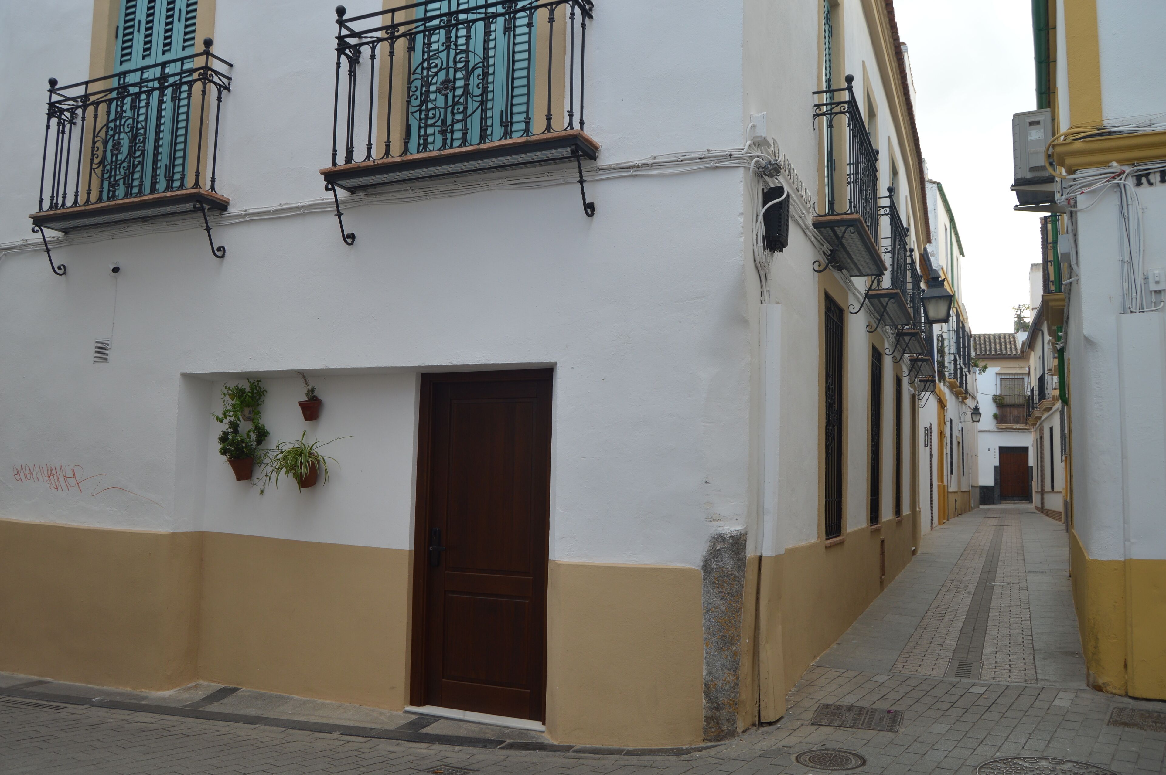A narrow street with buildings on either side, featuring balconies with metal railings and potted plants.
