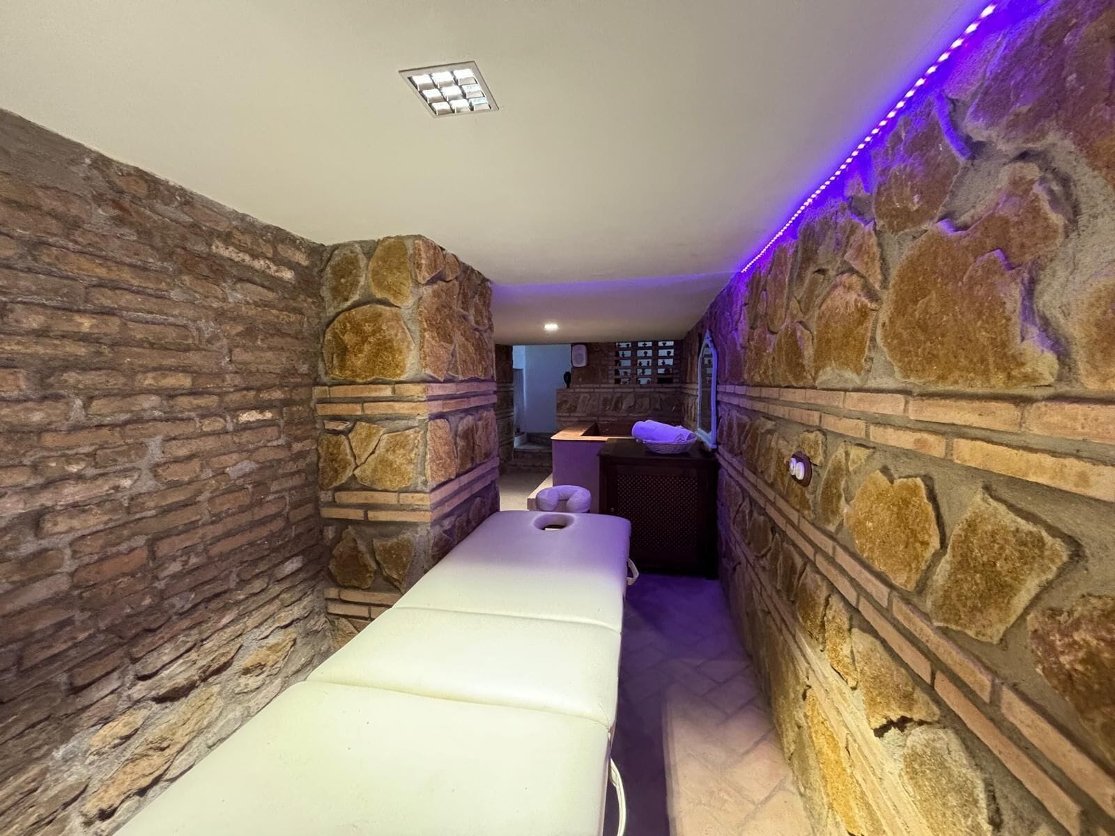 Een kamer met stenen muren, een massagetafel, een klein wastafeltje en een raam.