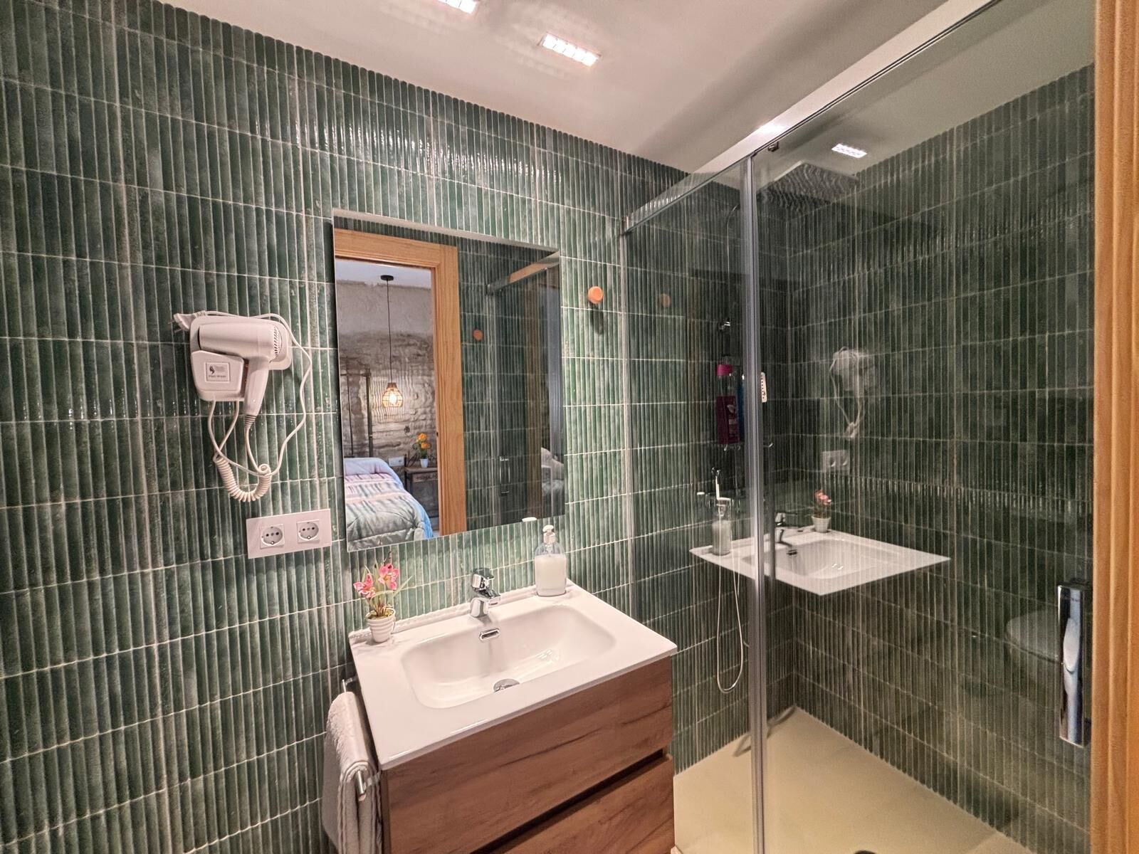 Ein Badezimmer mit grünen Fliesen an den Wänden, einem hölzernen Waschtisch, einem großen Spiegel und einer Duschkabine aus Glas.