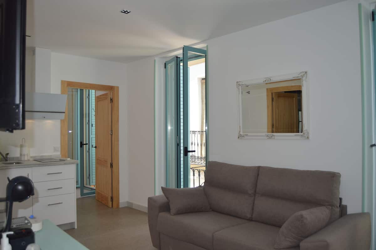 A living room with a beige sofa, a kitchen area with a white countertop, and a door leading to a balcony.