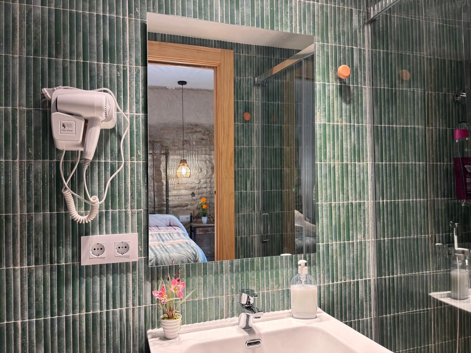 A bathroom with green tiled walls, a hairdryer on the wall, a mirror, a sink with a soap dispenser, and a potted plant.