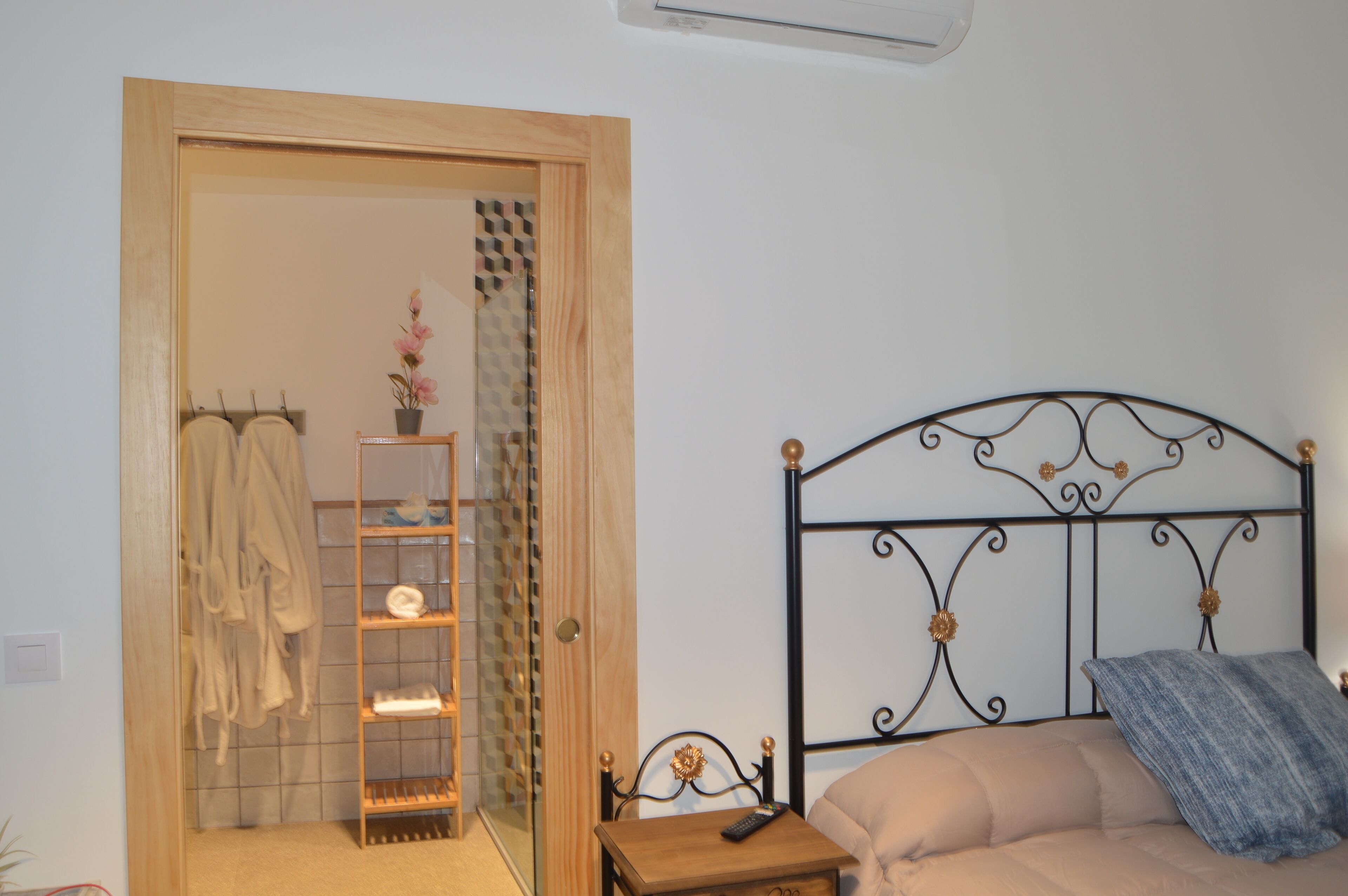 A bedroom with a bed, a wooden door, a shelf with towels, and a small table with a remote control.
